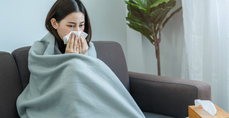 A young woman sitting on a sofa covered with a blanket, sneezing into a tissue due to flu or cold, illustrating symptoms of influenza and the importance of maintaining health and immunity